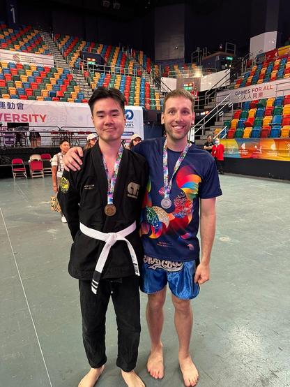 Picture of two people standing in a nearly empty stadium. On the left is an east Asian man wearing a black gi, white belt, and gold medal. On the right is a white masculine embracing him while wearing a Gay Games t-shirt, blue muay thai shorts, and a silver medal. Both are barefoot and smiling.