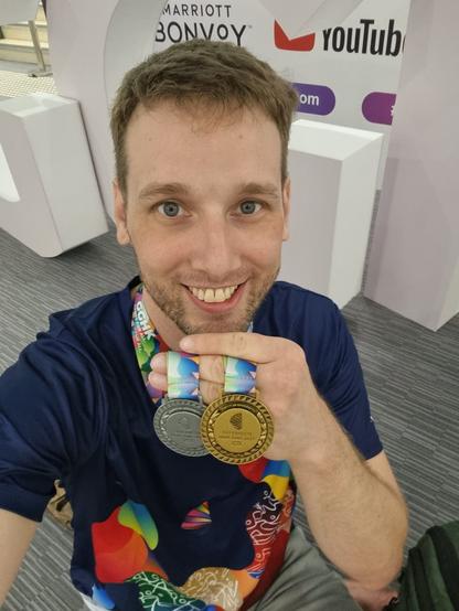 Shaun is a grinning white masculine person kneeling and holding up two Gay Games 11 medals, one silver and one gold. Both are engraved with Hong Kong symbols and Chinese characters. Shaun is also wearing a colourful Gay Games shirt.