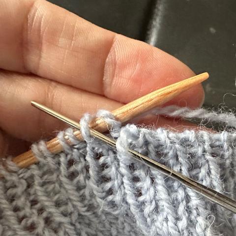 Light blue piece of knitting in 1x1 ribbing held in the left hand. The left stitches are on a wooden needle, the stitches on the right are bound off already. Through the last stitch on the right and the second stitch on the needle goes a darning needle.