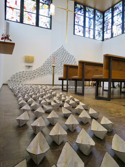 Der Innenraum der Klinikkapelle Rosenheim aus der Froschperspektive: Zu sehen ist eine Menge von Papierschiffchen mit dünnem schwarzen Rand, die sich von der Kamera weg Richtung Altar bewegen und dahinter die Wand hinauffahren; seine Spitze hat diese Flotte etwa auf der Höhe, auf der das von der Decke hängende goldglänzende Kreuz endet. Zu sehen sind auch die Bänke, die bunte Osterkerze auf einem Ständer und ein schlichter Tabernakel an der Wand. Nach oben hin schließen die weißen Wände des Raums mit großen, abstrakten Buntglasfenstern ab.