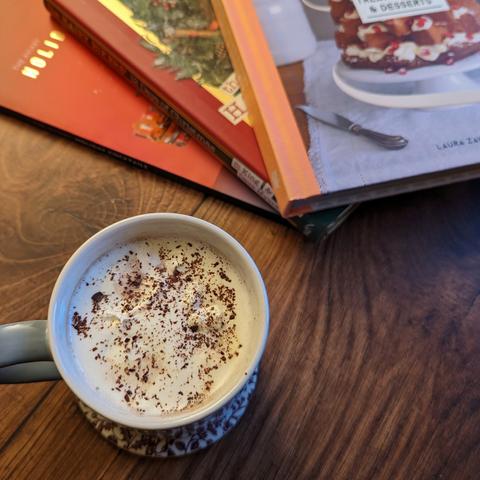 A mug with cream and chocolate sprinkles at the top, sitting beside some recipe books, on a dark wood background.