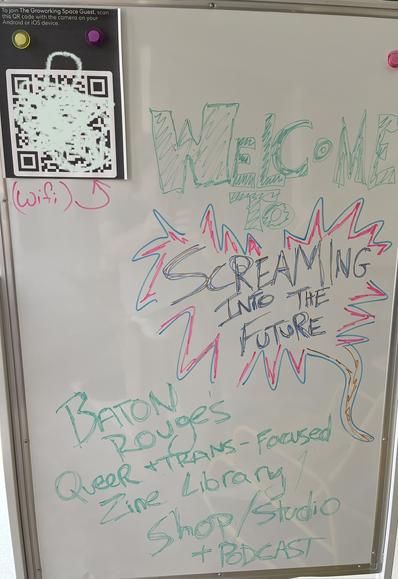 Dry erase board, “Welcome to Screaming Into The Future, Baton Rouge’s Queer and Trans Zine Library / Shop / Studio / Podcast