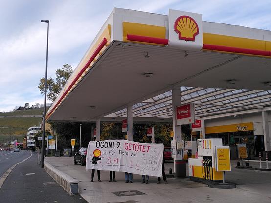 Bild einer Shell-Tankstelle, in der 5 Personen mit einem Banner stehen, auf dem "Ogoni 9 GETÖTET für Profit von SH_LL" steht.
