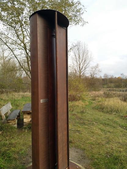 Eine 2,90 Meter hohe Säulenwindharfe aus Stahl auf einem Hügel in einer offenen Waldlandschaft platziert.