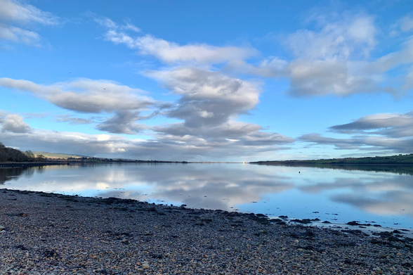 Cloud reflections in the cromarty firth. Blue sky silhouetted trees and land right and left. A greyed out pebble beach in the foreground