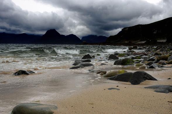 Ein bedrohlich wirkender schwarz-blauer Himmel über einer tiefschwarzen Bergkette. Darunter grau-grünes dunkles Wasser, Wellen und Gischt vom Sturm. Ein Strandabschnitt mit Felsen und Steinen.

A threatening black-blue sky over a deep black mountain range. Below, gray-green dark water, waves and spray from the storm. A section of beach with rocks and stones.