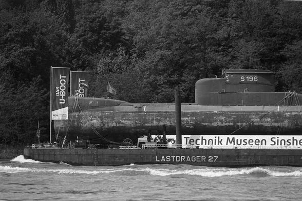 Schwarz-weiß: Der "Lastdrager 27" transportiert ein U-Boot über den Rhein. Ein Banner der "Technik Museen Sinsheim Speyer" ist zu sehen, ebenso zwei Fahnen am Bug mit der Aufschrift "Das U-Boot".