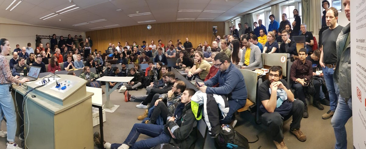 Photo of the Railways and Open Transport devroom at FOSDEM 2023. A room crowded with people who listen to a female speaker