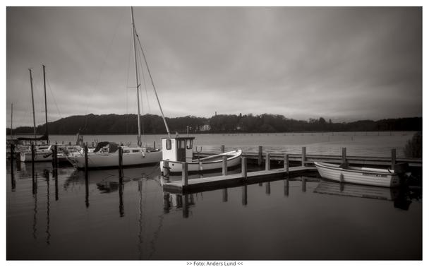 A black and white photo of some sailboats, a small fishing vessel and a motor dinghy at a dock, with a dramatic sky and trees with the white sanatory building amongst them in the background.