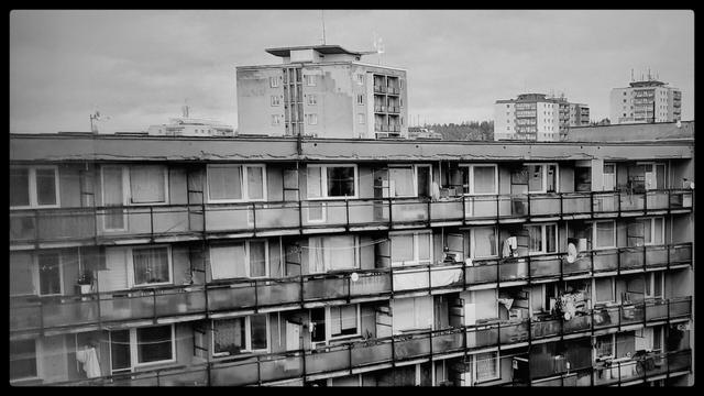 A dirty and damaged concrete block of small flats, small balconies side by side, a glimpse into the socialist history of the city. Black and white photograph.