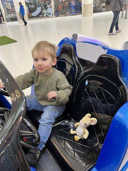 A blond haired toddler boy sitting in a blue plastic car in a shopping centre with his toy mouse carefully placed in the passenger seat beside him. The boy is wearing a khaki jumper and blue jeans.