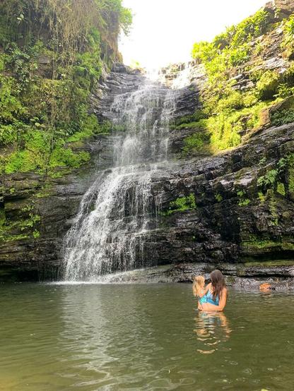A woman (me) holding a small girl stands in the pool of a beautiful waterfall.