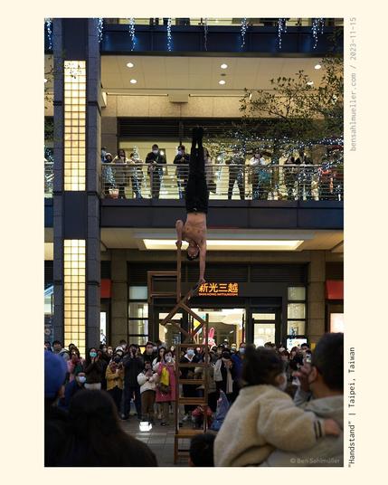 An athlete street performer doing a handstand on top of a pillar of chairs. A crowd gathered around him, watching in awe.