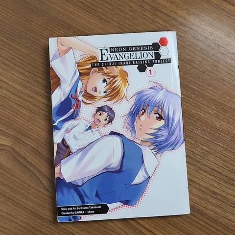 Photograph of the first volume of Neon Genesis Evangelion: The Shinji Ikari Raising Project sitting on a wooden desk. The cover has the name written on it along with Shinji, Rei, and Asuka all dressed in their school uniforms looking happy.