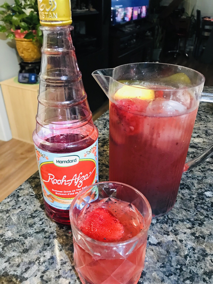 Bottle of Rooh Afza syrup on left; pitcher of sober sangria on right contains ice, lemon, strawberry, blueberry, sparkling water; glass of sangria with one strawberry in front of both! Pic by Crissy QTpi