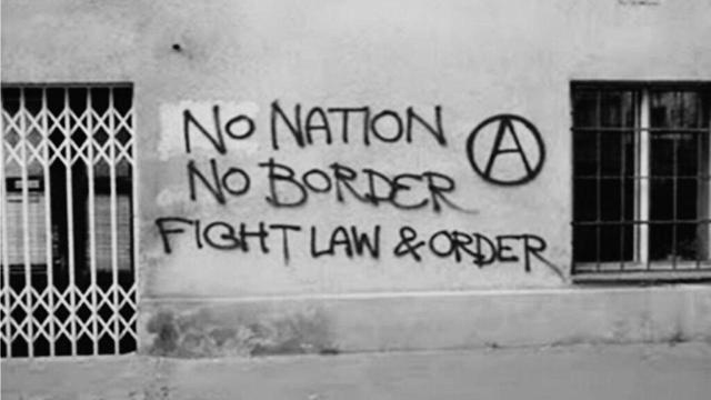 gray wall with a black graffiti "no nation no border figth law and order" next to an anarchy sign