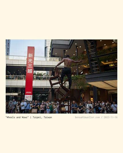 An athletic street performer standing victoriously on a pile of chairs. A crowd around him cheering him on excitedly.