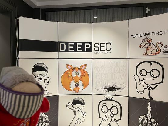 Foto of the grey sheep in front of a picture wall with the logos of the last several years of DeepSec.