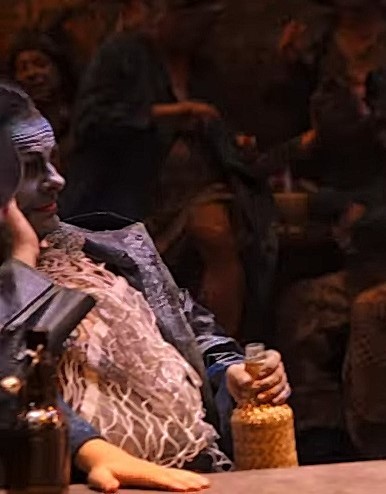 Dan as Bob Boles, seen right at the left edge of the photo, more so in profile. Mostly everyone had blue makeup for skin tones in the Erfurt production, looking striking against warm, orange toned backgrounds.

He's holding a jug-like bottle of booze, and wears a black leather coat + more visibly, a white fishnet style shirt of some kind, while sitting behind the pub table.

The old Twitter tag "Accidental renaissance" applies to the image or this crop of it. All photos are screen captures from trailers or other video footage.