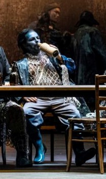 Bob Boles drinking from his bottle while manspreading behind the pub table. 

Dark hair, blue face, blue wellington boots on his feet, grey trousers and the white shirt of some kind made from literal fishnet looking things. 
The background is in the warmer brown tones, with 2 indistinct human figures further in the back facing each other.

Yes, I left the preview to show more of the crotch instead of the face. I have three other pictures for that, what he exudes here more this picture's thing.
