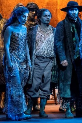 Fisher village chic. Where's our Innsmouth again?

A striking looking long haired woman next to two men, the one on the right wearing black clothes + a hat. All with blue makeup all over whatever skin + hair visible. Fishnets for clothes to some extent at least. Dan's Bob Boles in the middle.

You get the impression that the people who had offspring with the fish people "Deep Ones" in Innsmouth were not that far off from the vibes here. H.P. Lovecraft story reference.

The background is aflame in orange colour contrasting against the blue tones of the people.