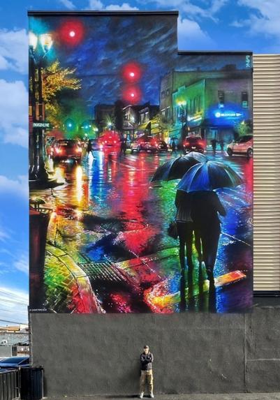 Streetartwall. This fantastic mural of a city scene in the rain and at night was sprayed/painted on the exterior wall of a multi-storey building. The manga-style scene of the shopping street "Bergenline Avenue" with neon signs, streets, cars and evening sky looks very lively and is fantastically realized. A few cars drive quickly through the colorful streets and two passers-by stand on the street under umbrellas in the foreground. The lanterns and traffic lights are illuminated and the scenery is reflected on the rain-soaked asphalt.