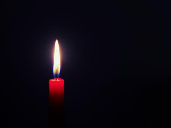 A burning, red candle on a very dark background with a hint of blue. The light of the candle is only illuminating the top part the the candle. The rest is in the dark. The flame is burning blue at the very bottom and is bright orange for the rest of the flame. Around the middle of the flame there is kind of a red halo.