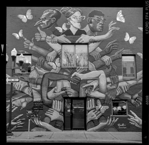 Black and white photo of a building entrance that has many windows over two floors and all of it is tied together with a mural on the brick wall. The mural is three figures and a lot of different hands and arms holding each other.