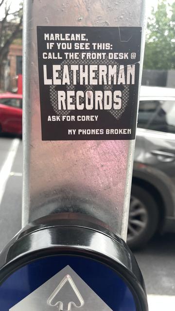 A cheap home made looking graphic designed sticker on the pole at a pedestrian crossing, bold white text on black reads 

MARLEANE,
IF YOU SEE THIS:
CALL THE FRONT DESK @ LEATHERMAN
RECORDS
ASK FOR COREY
MY PHONES BROKEN