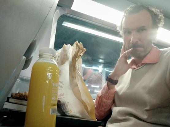 A bottle of orange juice, a bag with pastries, and a serious looking handsome middle aged man seating on a TGV train