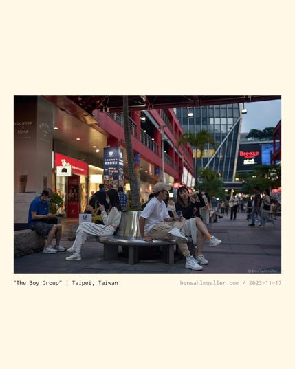 Shoppers relaxing on a bench in the street, listening to a small boy band.