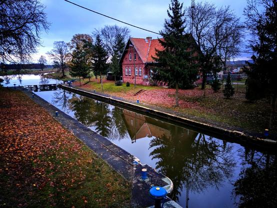 Śluza na Noteci pełna wody. Na przeciwległym brzegu dom z czerwonej cegły kryty dachówką otoczony drzewami, odbijający się w lustrze wody.