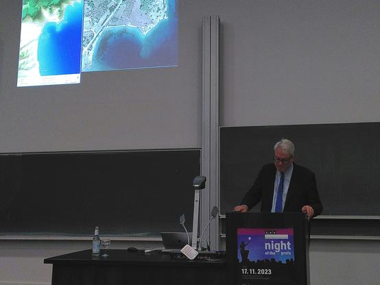 A man stands at a lecturn in a lecture hall. A poster displays the words Night of the Profs. Satellite images of a coastline are displayed at the wall behind the.man.