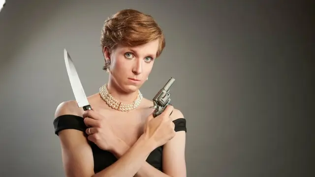 Linus Karp, a thin white man with light brown hair, portrays Princess Diana of Wales, in a strappy black, low necked dress (a.k.a the Revenge Dress) with a three layered pearl necklace, holding his arms crossed over his chest, a chef's knife in one hand, and a pistol in the other.