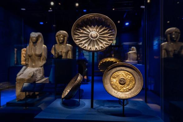 Against a deep blue background, a view from the Ramses exhibition shows golden shields displayed in the foreground, while behind them are shown a number of life-size seated figures of the Ramses family and gods, all carved in stone.