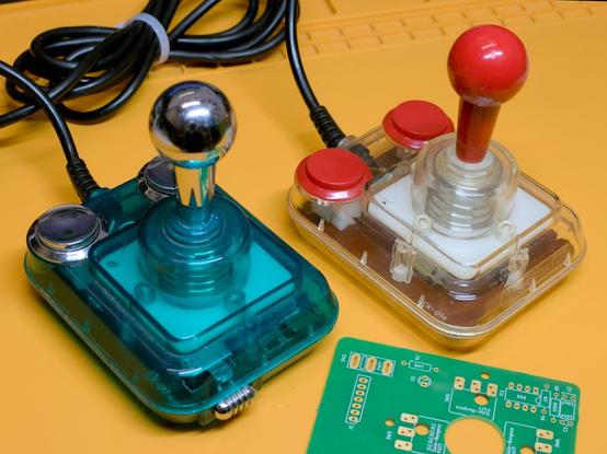 On the left: A green Competition Pro Mini joystick with silver stick and silver buttons. On the right: A clear Competition Pro Mini with red stick and red buttons. At the front there is a corner of an unpopulated PCB.