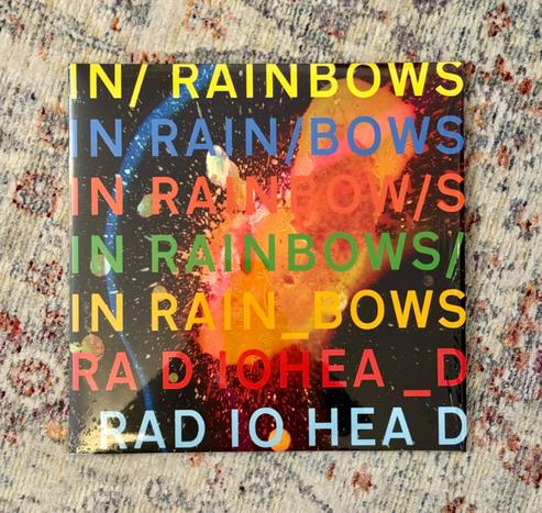 Photo of a record album cover laying on a pale rug. 

The cover is a splash of colours against a black background with multicoloured text reading, ‘In Rainbows’, and ‘Radiohead’.