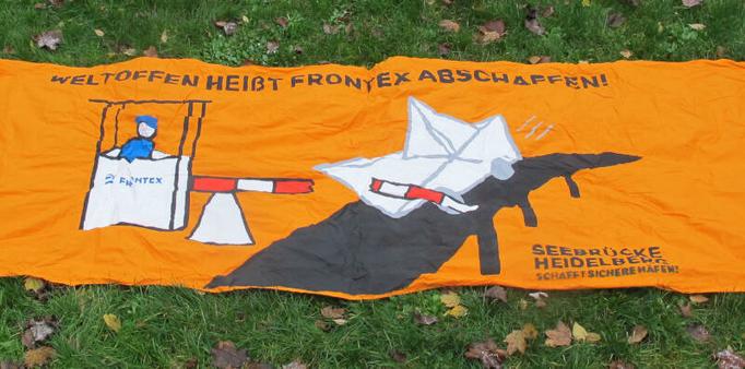 Oranges Transparent auf grüner Wiese: „Weltoffen heißt Frontex abschaffen“ und ein ein Papierschiff, das einen Schlagbaum durchbricht.