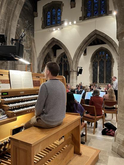 An organist sitting at the console, with an orchestra preparing to start rehearsing in the background. All in an old church with arches etc.