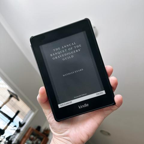 A hand holds a Kindle showing the cover of Mathias Enard’s THE ANNUAL BANQUET OF THE GRAVEDIGGERS’ GUILD.
