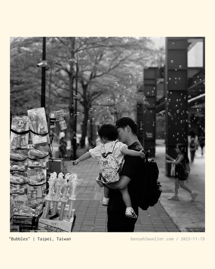 A young girl carried on her mother's arms, excitedly pointing at some bubble-making-machine.