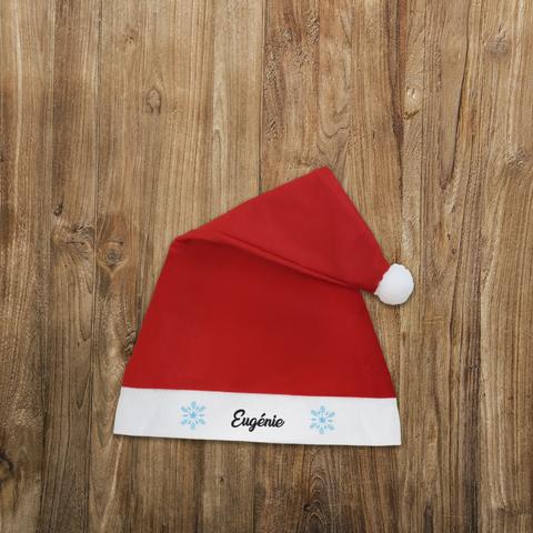 Bonnet de Noël rouge avec un pompon blanc et personnalisé avec le prénom de votre choix entouré de flocons de neige. A retrouver dans notre boutique en ligne : ernestine-etcie.fr