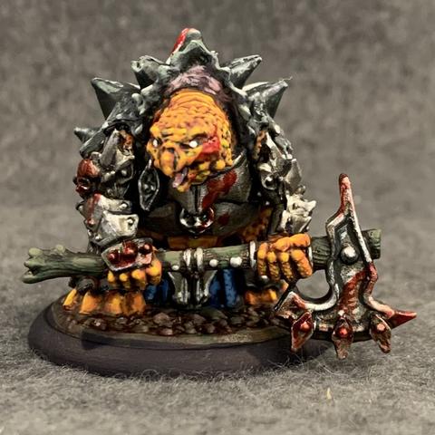 Stout bipedal turtle-person in heavy plate armor and a giant black shell with a white skull painted on the back. He holds a giant blood-smeared two handed axe. He has rough orange skin, and he glares with pupil-less eyes and an open mouth. Blood is also smeared across his face, armor, and shell.