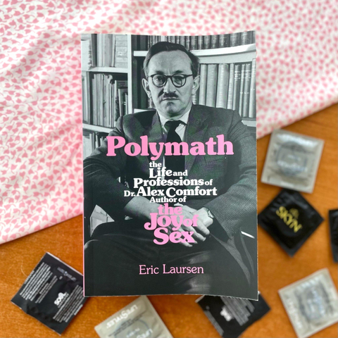 A copy of "Polymath" from AK Press sits on top of a table draped in a baby pink scarf. The cover of the book is a black and white photo portrait of Alex Comfort with pink text. Next to the book on the table are an assortment of condoms in grey and black wrappers.