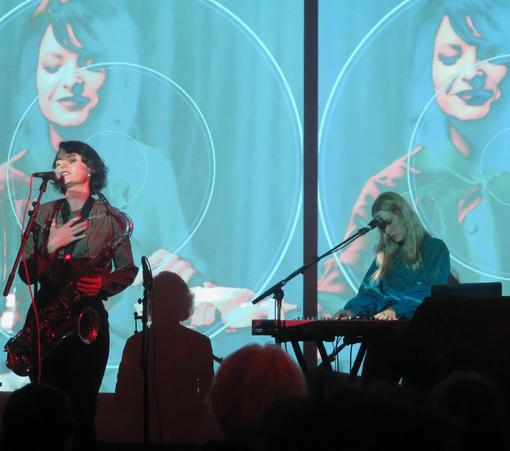 Two women performing onstage, with the backs of audience heads in front of them and a projection of another woman onto a screen behin them. One of the women is seated and playing keyboards, while the other is standing and seems to be singing into a microphone but is also holding a saxophone.