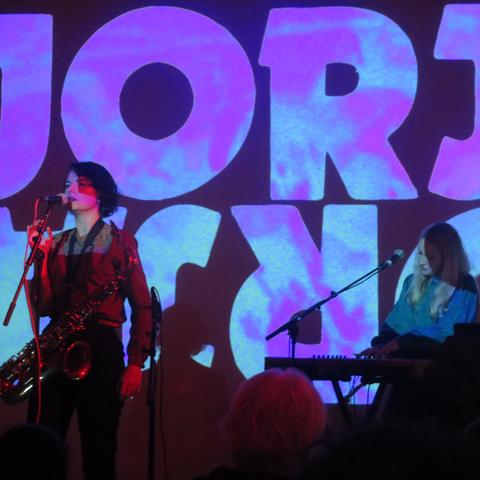A darker image showing the same two women performing onstage as in the previous picture. This time there are letters being projected onto the screen behind them, which look like they might be saying JORJA twice, the second time backwards underneath the first.