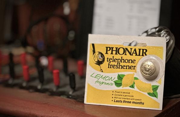 Manual telephone switchboard key shelf, displaying a 2β square yellow cardboard blister pack, with a small metallic disc in the plastic blister.
Packaging illustration shows a GPO 700 style handset with the metallic disc clipped into the holes on the mouthpiece.
Advertising/packaging text reads:
Phonair telephone freshener
Lemon fragrance
* fixed in seconds
* contains a germicide
* will not interfere with speech
* lasts 3 months