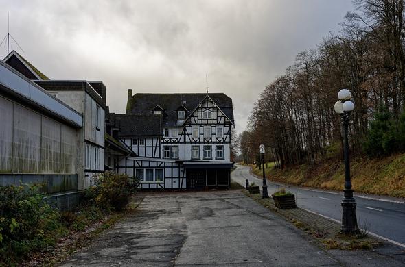 Links ein riesiger verfallender Fachwerkhauskomplex. Im Vordergrund links ein Parkstreifen. Halbrechts zieht sich die Hauptstraße von unten nach oben durch das Bild. Rechts Bäume am Straßenrand.