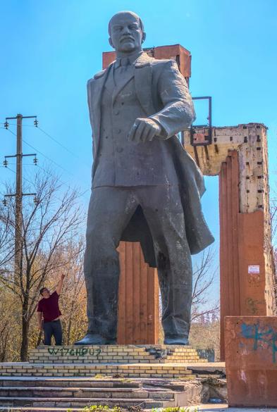 A huge statue of Vladimir Lenin towers over a man pointing upwards. The man only comes up to Lenin's shins.