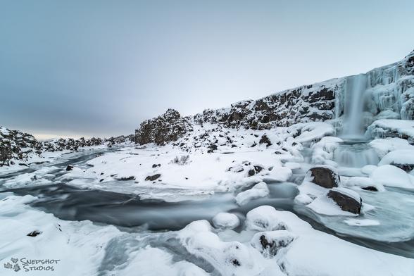 Wide angle view of a narrow valley covered by snow and ice. A waterfall comes down the cliff on the right, and the creek goes down the valley on the left.